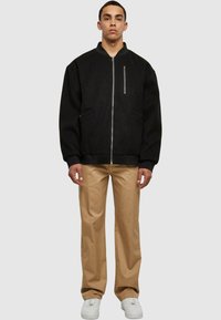 Chaqueta negra con cremallera y cuello alto, con dos bolsillos laterales y un bolsillo en el pecho; combinada con pantalones beige de pierna recta y zapatillas blancas.