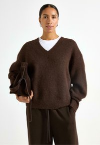 Donna che indossa un maglione fuzzy marrone con scollo a V e pantaloni abbinati, mentre tiene una borsa di camoscio marrone scuro su uno sfondo bianco.