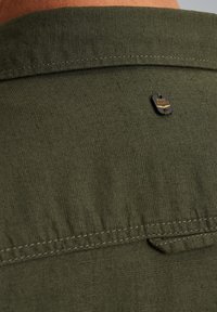 Detalle de camisa de tela verde con textura tejida, costuras visibles y una etiqueta de marca con el logo "PME LEGEND" en la parte posterior.