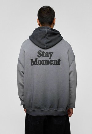 Hoodie in een verloop van grijs, met een zachte textuur. Heeft een gedurfde "Stay Moment" print op de achterkant en ribbels aan de mouwen en onderkant.