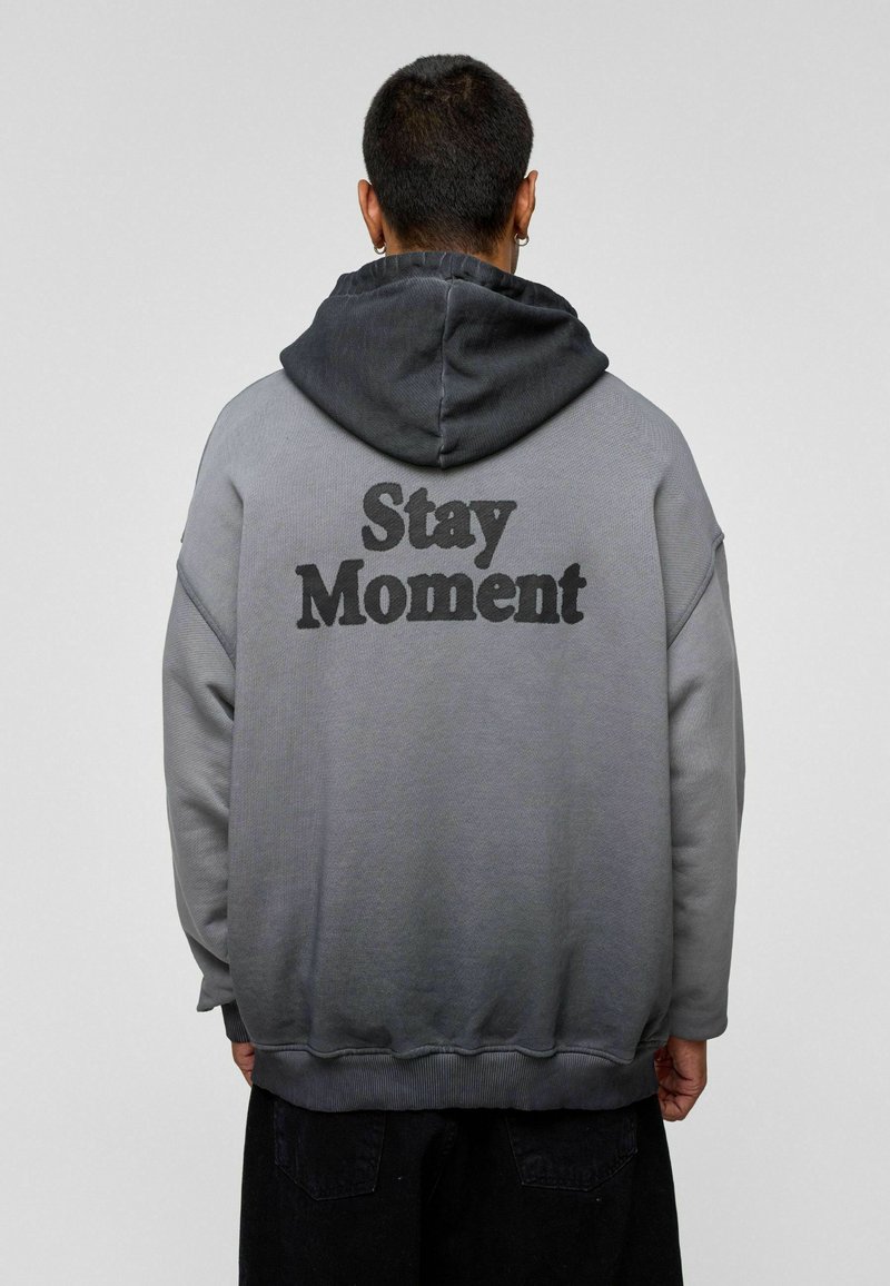 Hoodie in een verloop van grijs, met een zachte textuur. Heeft een gedurfde "Stay Moment" print op de achterkant en ribbels aan de mouwen en onderkant.