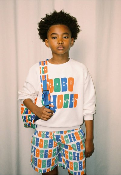Sweatshirt blanc avec le texte coloré "BOBO CHOSES", shorts assortis, et un sac à épaule à motifs avec des sangles bleues. Tissu texturé.