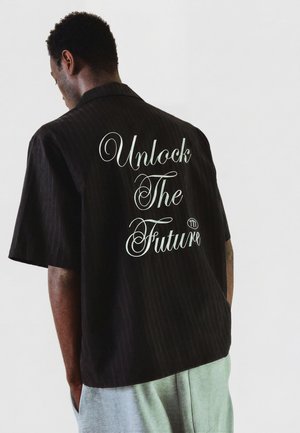 Férfi háttal, fekete rövid ujjú pólót visel, amelynek hátán fehér kurzív szöveg: „Unlock The Future”.