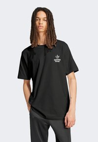 Black cotton t-shirt with a relaxed fit, featuring a white "superstar PARIS" logo and an iconic three-stripe emblem on the chest.