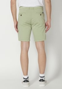 Pantalones cortos de algodón de color verde claro con corte recto, dos bolsillos traseros y detalles de botones marrones, combinados con calcetines a rayas blancas y negras y zapatillas deportivas.