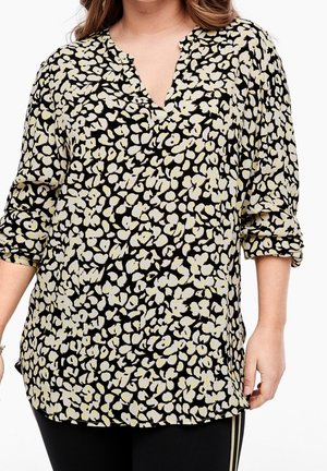 Femme portant un chemisier noir ample avec un imprimé floral abstrait crème et jaune pâle, manches retroussées, vue du cou jusqu'aux hanches.