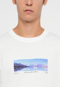 Detailní záběr na hruď osoby v bílém tričku s modrou a purpurovou abstraktní krajinou a kurzívou textem pod ním.