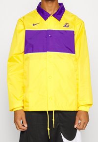 Hombre vistiendo una chaqueta de chándal amarilla y morada de los Lakers con el logotipo de Nike, combinada con pantalones cortos negros y blancos de Nike, de pie con las manos a los lados.