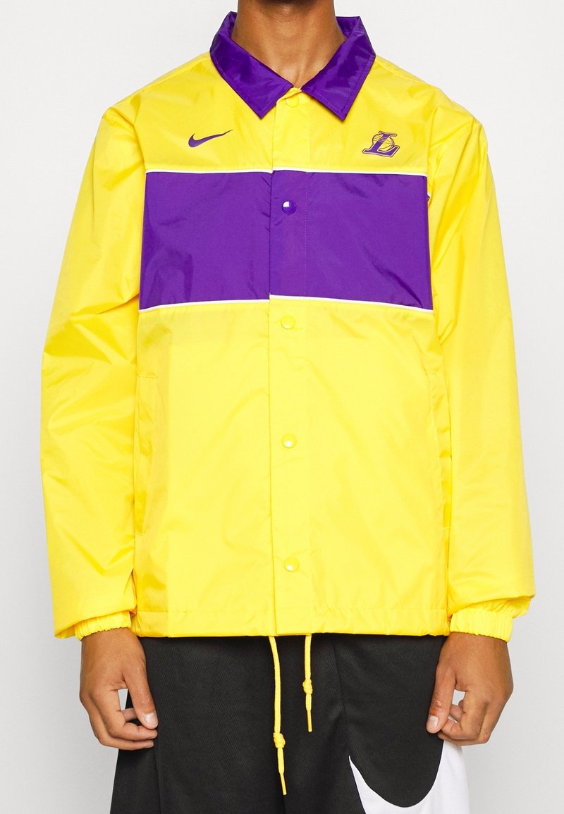 Hombre vistiendo una chaqueta de chándal amarilla y morada de los Lakers con el logotipo de Nike, combinada con pantalones cortos negros y blancos de Nike, de pie con las manos a los lados.