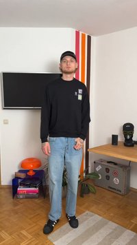 Melna sporta krekls ar logo, gaiši zilas džinses un melnas kedas ar baltām svītrām. Stāv istabā ar krāsainiem akcentiem un mēbelēm.