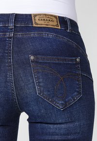 Jeans de denim azul oscuro con un diseño distintivo de costura en los bolsillos curvados. Presenta una etiqueta de cuero en la cinturilla y detalles metálicos visibles.