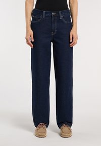 Persona che indossa jeans dritti blu scuro e scarpe casual beige con lacci, in piedi contro uno sfondo bianco semplice.