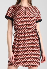 Kurzärmeliges Kleid mit einem roten und cremerfarbenen dreieckigen Muster. Elastischer Taillenbund mit Kordelzügen und kontrastierenden schwarzen Akzenten.