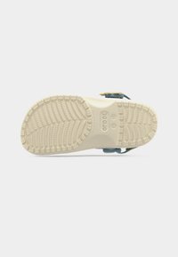 Semelle d'une sandale Crocs beige avec une bande de roulement texturée et une bride visible sur le côté, présentée sur un fond blanc uni.