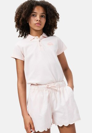 Niña con cabello rizado, vistiendo una camiseta tipo polo rosa claro y pantalones cortos a juego con un borde ondulado, de pie con una mano en el bolsillo.