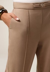 Pantalons beige ajustés avec une texture lisse, des poches latérales et une taille étroite avec un détail de boucle dorée.