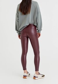 Person som står och bär burgundyröda fusk läderleggings, en ljusgrå överdimensionerad sweatshirt, och vita sneakers med lila och olivfärgade detaljer.