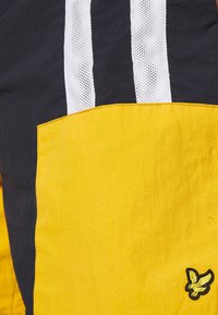 La sezione della giacca nera e gialla presenta una grande tasca gialla, strisce in rete bianche e un piccolo logo giallo ricamato sulla tasca.