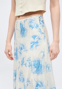 Γυναίκα που φοράει μπεζ cropped τοπ και φούστα με ψηλή μέση σε λευκό χρώμα με μπλε λουλουδένιο σχέδιο και φερμουάρ στο πλάι.