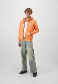 Narancssárga zipzáras kapucnis pulóver kenguruzsebekkel és fehér grafikai szöveggel, világoskék, bő szárú farmernadrággal és fekete cipővel párosítva.