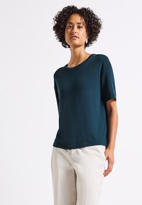 Top corto colore teal scuro con scollatura rotonda, caratterizzato da una vestibilità morbida e una texture liscia. Abbinato a pantaloni beige chiaro.