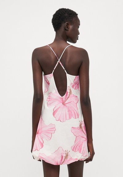 Vestido floral con patrones de hibisco rosa sobre una tela blanca. Presenta un corte en la espalda bajo y delgados tirantes entrelazados. Textura suave.