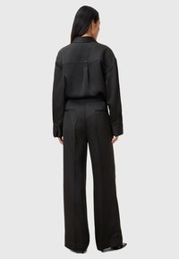 Camicia nera su misura e pantaloni a gamba larga realizzati in tessuto morbido; presenta uno spacco posteriore e dettagli discreti.