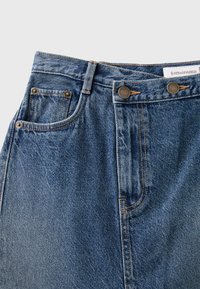 Jean en denim bleu en gros plan montrant la poche avant, la boucle de ceinture, la fermeture à double bouton et l’étiquette de la marque à l’intérieur de la ceinture.