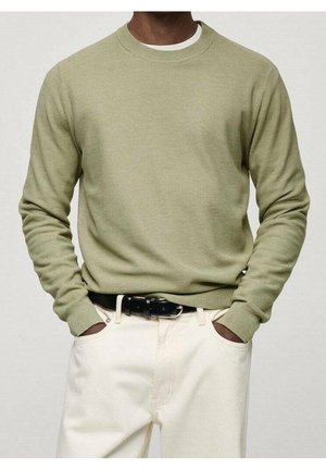 Person iført en lysegrøn crewneck-trøje, hvid undertrøje, hvide bukser og sort bælte med hænderne i lommerne.
