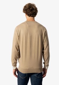 Suéter de punto beige con mangas largas, cuello redondo y puños de marca blancos. Textura suave con una silueta ajustada. Combinado con jeans azules.
