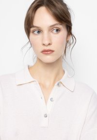 Maglione polo in maglia beige chiaro con colletto, tre bottoni grigio chiaro e una texture morbida. Design semplice senza motivi visibili.