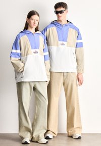 Világoskék, bézs és fehér színblokkolt, kapucnis szélálló kabát első logóval, laza pasztellszínű nadrággal párosítva. Texturált anyag, sportos kialakítás.