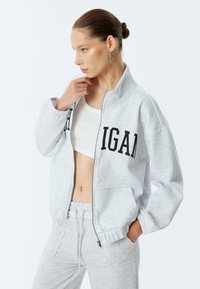 Sudadera de cremallera color gris claro con un alto cuello, letras negras en la parte frontal y tejido texturizado. Combinada con pantalones de chándal a juego.
