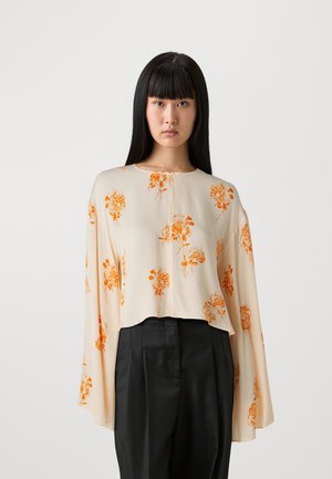 Kremfarget bluse med oransje blomstertrykk, lange, vide ermer og rund halslinning. Sammen med svarte bukser for kontrast.