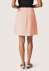 Korallo spalvos A formos sijonas su lygia tekstūra, iki vidurinės šlaunies ilgio ir dviem užpakalinėmis klostėmis. Su juodomis aukštakulnėmis šlepetėmis.