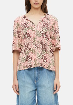 Frau trägt eine kurzärmelige Bluse mit rosa-braunem Blumenmuster, kombiniert mit hellblauen hoch taillierten Jeansshorts.