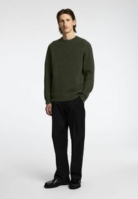 Suéter ribeteado color verde oliva con cuello redondo, llevado con pantalones negros y zapatos negros. La tela tiene una apariencia texturizada, ofreciendo un ajuste relajado.