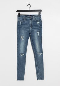 Hubené džíny ve středně modrém denimu s děravými místy, třepeným lemem a klasickým pětipocket designem, visící na černém ramínku.