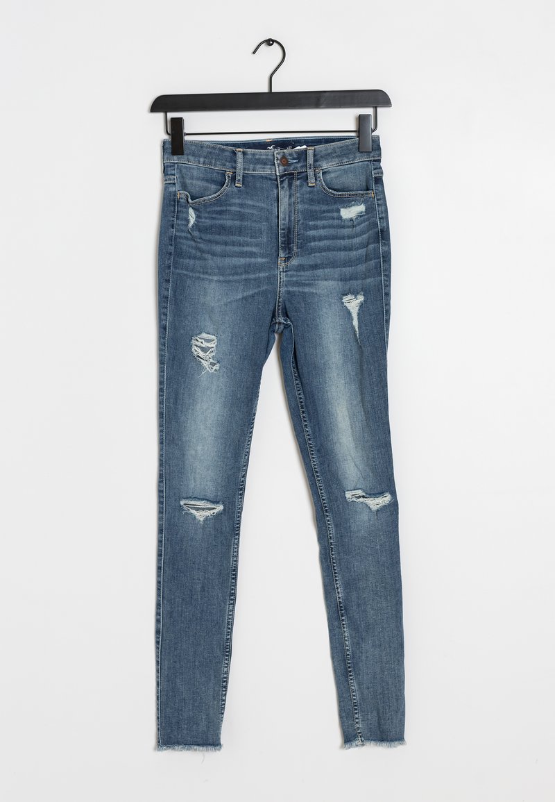 Hubené džíny ve středně modrém denimu s děravými místy, třepeným lemem a klasickým pětipocket designem, visící na černém ramínku.