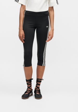 Άτομο που φοράει μαύρα κοντά κολάν με λευκές πλαϊνές ρίγες και λογότυπο Adidas, σε συνδυασμό με μαύρα μπαλαρίνες με κορδόνια που τυλίγονται γύρω από τον αστράγαλο.