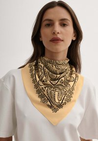 Jedwabny szalik w jasnym żółtym kolorze z czarnym wzorem paisley, w kształcie trójkąta, o teksturowanym wykończeniu. Noszony na szyi na białej koszuli.