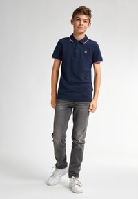 Námořnická polokošile s límcem v červenobílých pruzích, s knoflíkovou légu na tři knoflíky, nošena s šedými džíny a bílými teniskami, stojící na šedém pozadí.