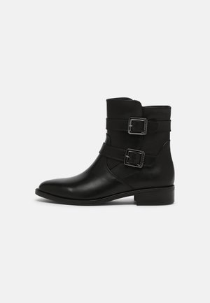 Bottine en cuir noir avec deux sangles argentées bouclées et un petit talon sur fond uni gris clair.