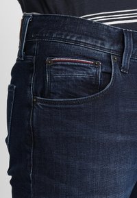 Jeans in denim blu scuro con una vista ravvicinata della tasca anteriore, caratterizzati da cuciture rosse e un tessuto leggermente testurizzato.