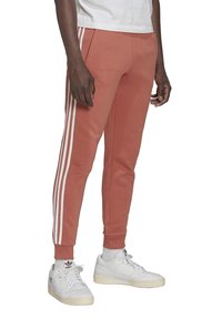 Red-orange athletic joggers featuring three white side stripes, tapered legs, and a comfortable fit paired with white sneakers.