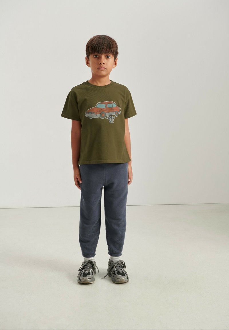 Camiseta verde oliva con gráfico de coche naranja, combinada con pantalones deportivos azul marino ajustados y zapatillas grises, sobre un fondo neutro.