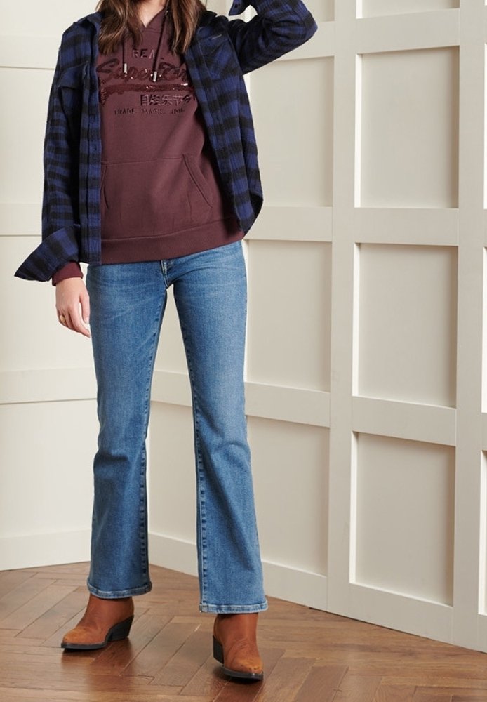 Femme portant un sweat à capuche marron et une chemise à carreaux bleue par-dessus un jean bleu et des bottines marron, debout sur un sol en bois.