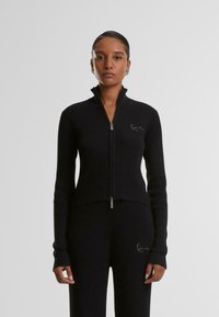 Pull à zip noir côtelé avec un col haut et des manches longues, présentant un petit logo brodé sur la poitrine gauche et une coupe ajustée.