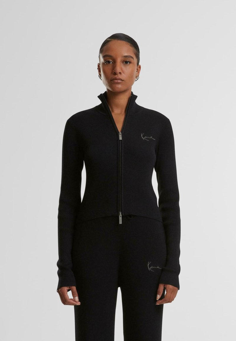 Pull à zip noir côtelé avec un col haut et des manches longues, présentant un petit logo brodé sur la poitrine gauche et une coupe ajustée.