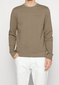 Sudadera de algodón marrón con mangas largas y cuello redondo. Presenta un sutil logotipo en el pecho y puños y dobladillo de canalé. Combina con pantalones blancos.